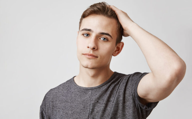 Seriously looking young handsome man with blue eyes touching his hair. Day was hard for this person so he looks tired, thinking about time he will finally come home and take a nap.